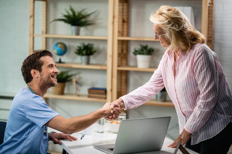 A smiling male healthcare professional in scrubs shakes hands with an older woman in a striped shirt across a desk with a laptop, in a bright, modern office setting.