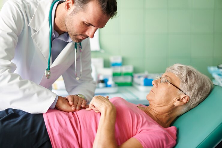 A doctor examines an older woman lying on a medical bed, gently pressing on her abdomen. The woman looks up at the doctor while in a clinical setting with medical supplies in the background.