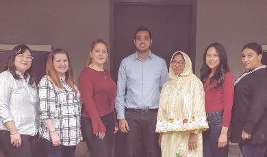 About Us Photo Seven people stand in a line indoors, posing for a group photo. Most are smiling at the camera. The group includes women in business-casual attire and one wearing a light-colored traditional outfit and headscarf.