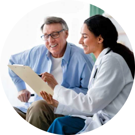 ForProvidersAssets_photo2 A smiling doctor in a white coat sits beside an older man, showing him a clipboard. The man is also smiling and holding a pen, appearing engaged in the conversation.