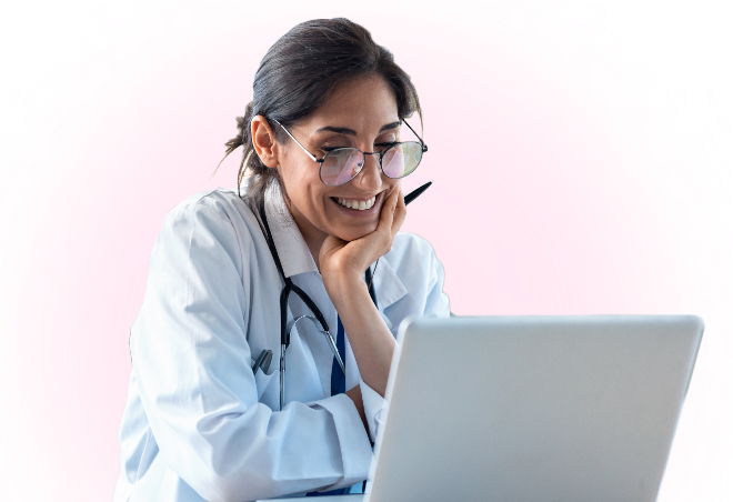 Mask Group 3 A smiling doctor wearing glasses and a white coat with a stethoscope looks at a laptop, resting her chin on her hand, in front of a pink background.