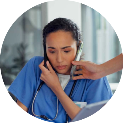 Partner_Photo3 A nurse in blue scrubs looks stressed while holding a phone to her ear with her shoulder, as another hand offers her a second phone. She appears overwhelmed and busy in a hospital setting.