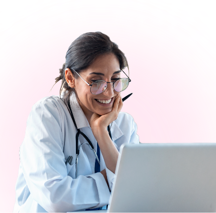 Photo A smiling doctor wearing glasses and a white coat with a stethoscope looks at a laptop, sitting against a pink background.