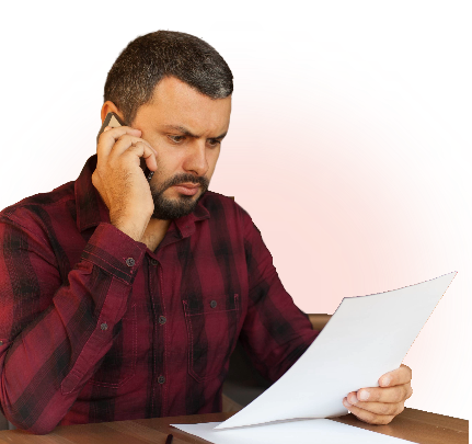A man with short dark hair and a beard is sitting at a table, talking on a smartphone while looking seriously at a sheet of paper he is holding in his other hand.